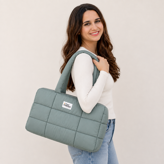 A woman with long brown hair smiling and holding a large quilted sage green tote bag over her shoulder, wearing a white long-sleeve top and light blue jeans against a plain beige background.