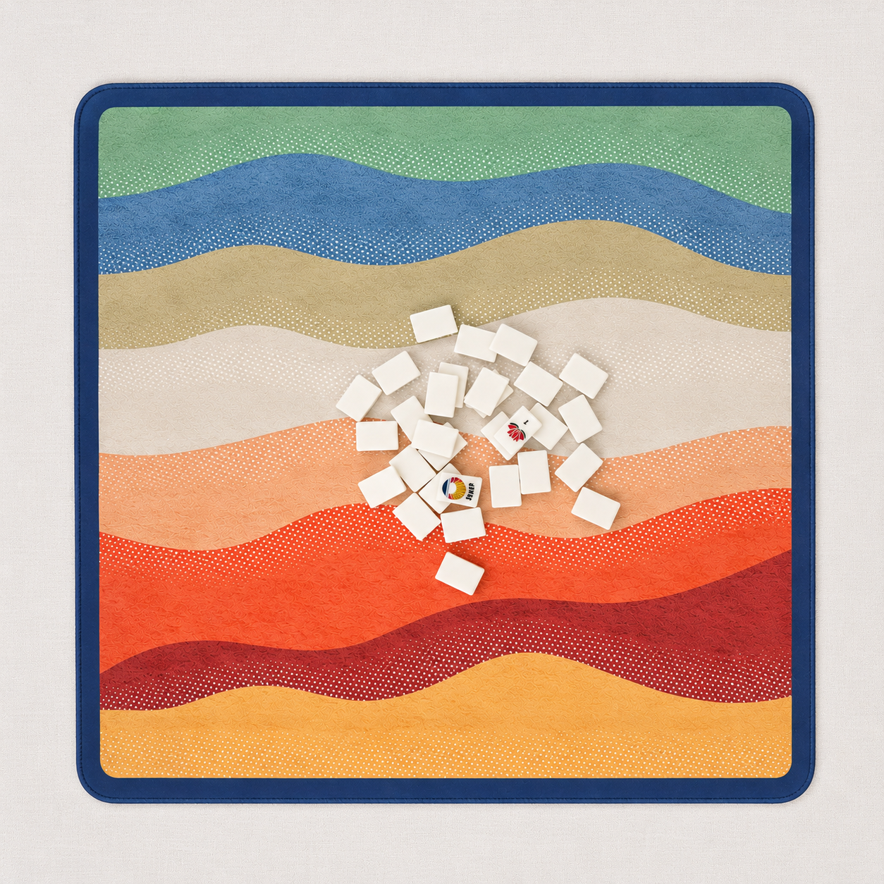 A square neoprene mahjong mat with rounded corners, featuring wavy stripes in orange, red, peach, cream, tan, blue, and green on a dark navy border. White rectangular travel size mahjong tiles are scattered near the center of the mat on a light neutral background.
