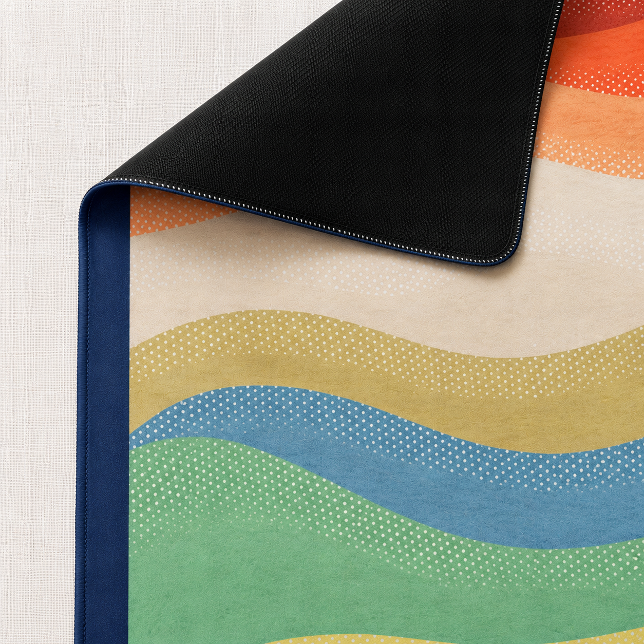 A close-up of a colorful mahjong mat with a wavy, dotted pattern in green, blue, yellow, orange, and beige, shown with one corner folded over to reveal a black neoprene textured underside and a navy blue edge.