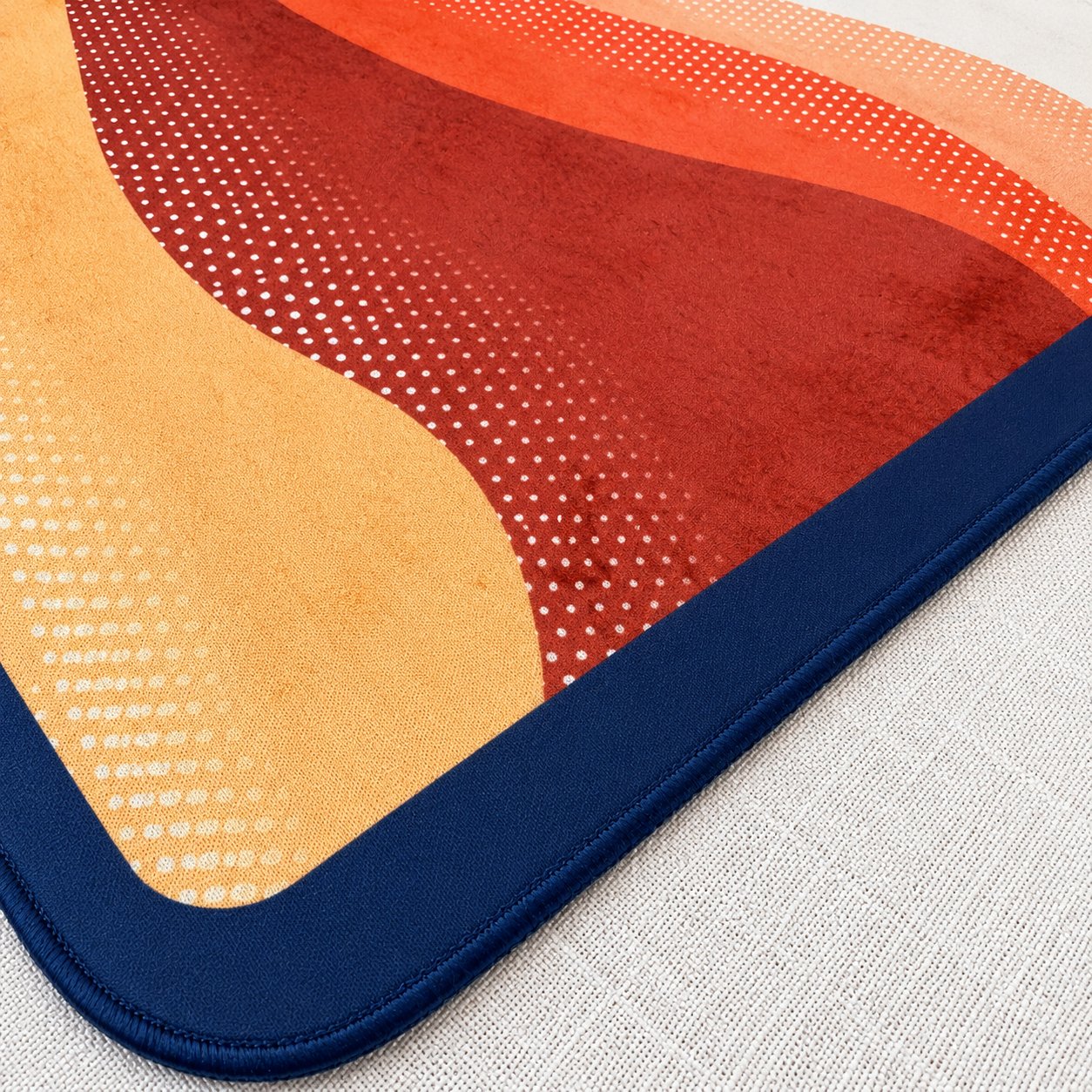 A close-up of a square mahjong mat corner showing a wavy abstract pattern in beige, orange, red, and navy blue, with a thick navy border with matching navy stitching on a light textured surface.